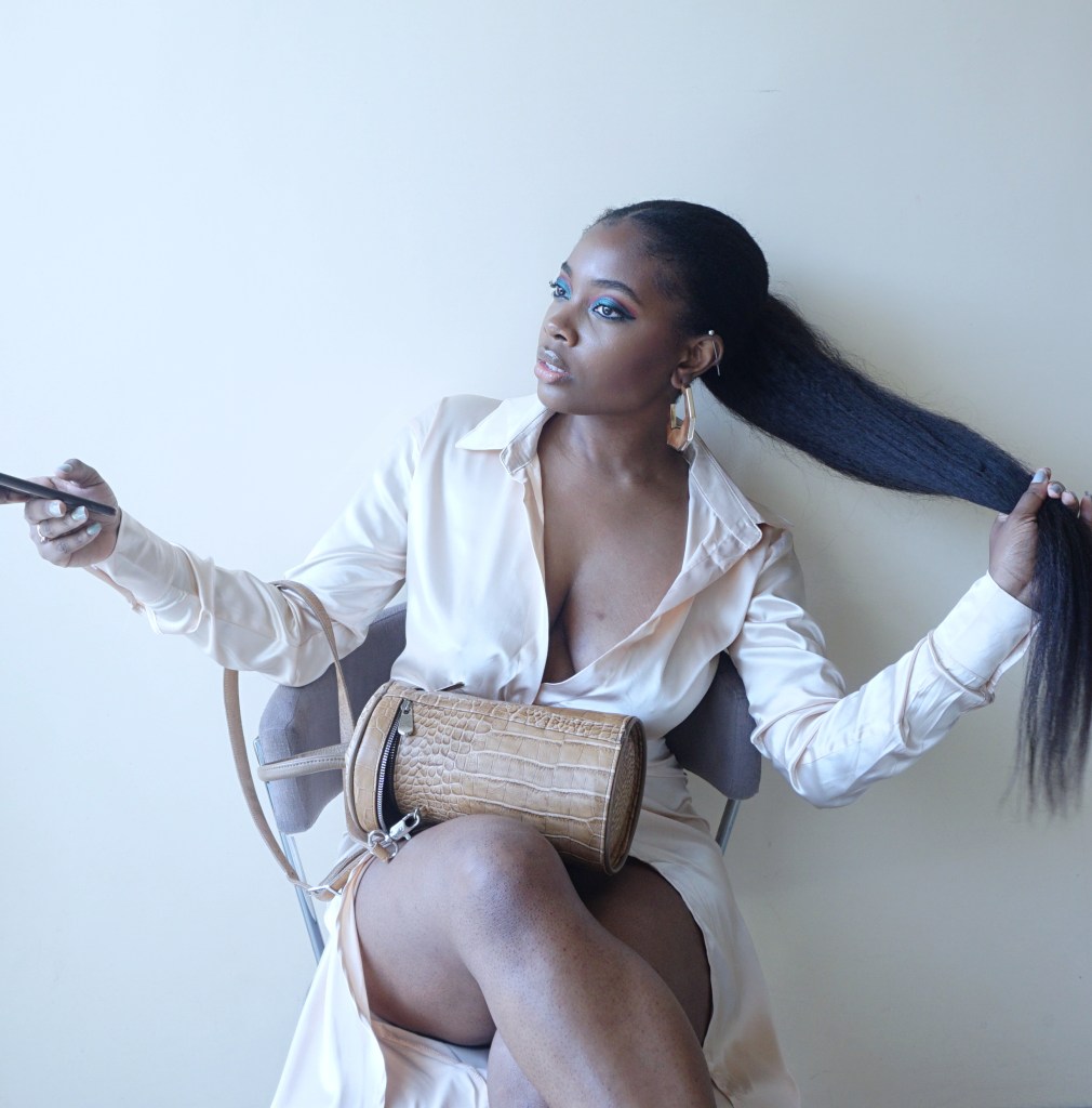 Woman sitting on chair with leather bag on her lap, holding her hair in one hand and her phone in the other.