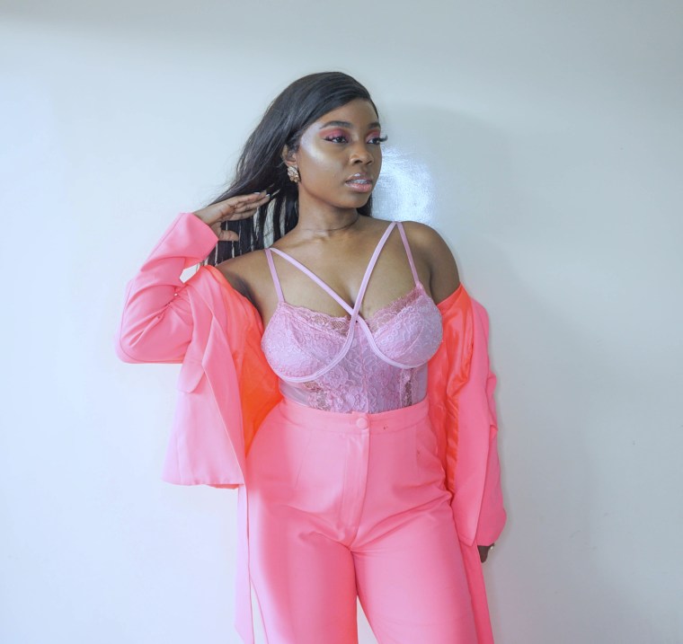 Head to knee shot of a woman standing in front of a plain background with one hand in her hair