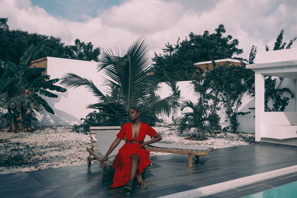 Block woman sitting in the backyard of a home, wearing a long red dress with her legs crossed.