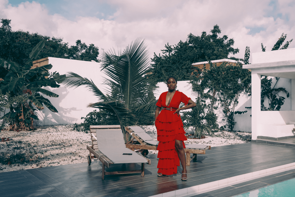 Woman standing in the backyard of a home with palm trees, in a long red dress and one hand on her waist