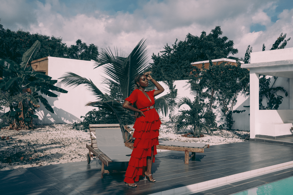 Woman standing sideways in the backyard of a home, near a pool, looking away with her head up, while wearing a long red dress